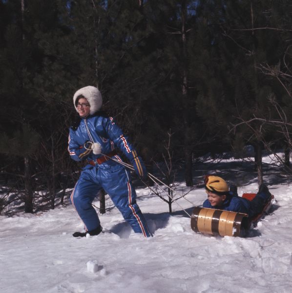 A young woman is pulling a a boy laying in a toboggan across the snow. Trees are behind them.