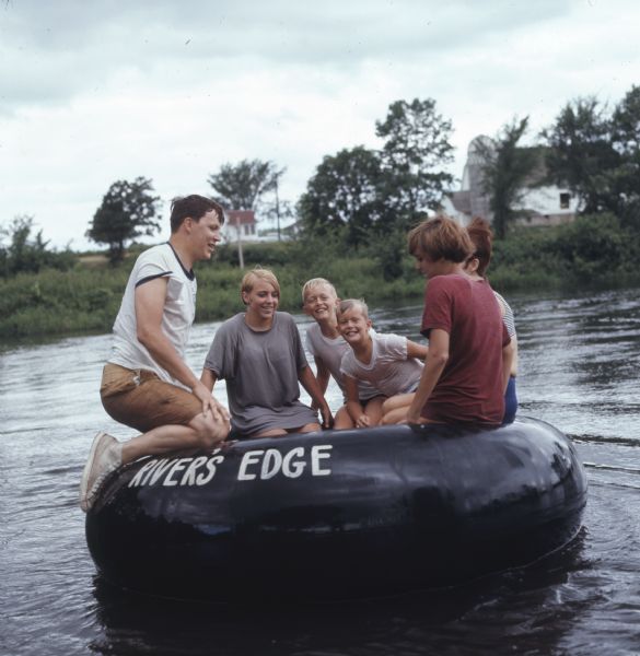A group to children and young adults are sitting on a large rubber inner tube on the Apple River. "River's Edge" is painted on the side of the tube. On the far shoreline are farm buildings.
