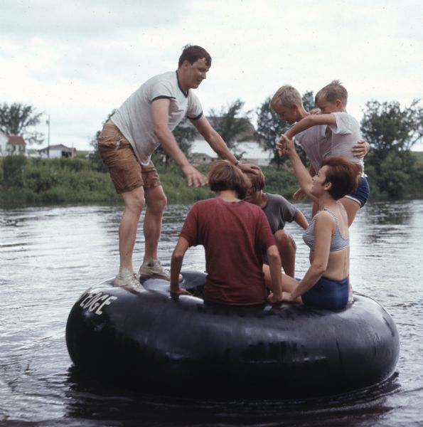 A woman and a group of children are playing on a large rubber inner tube on the Apple River. "River's Edge" is painted on the side of the tube. On the far shoreline are farm buildings.
