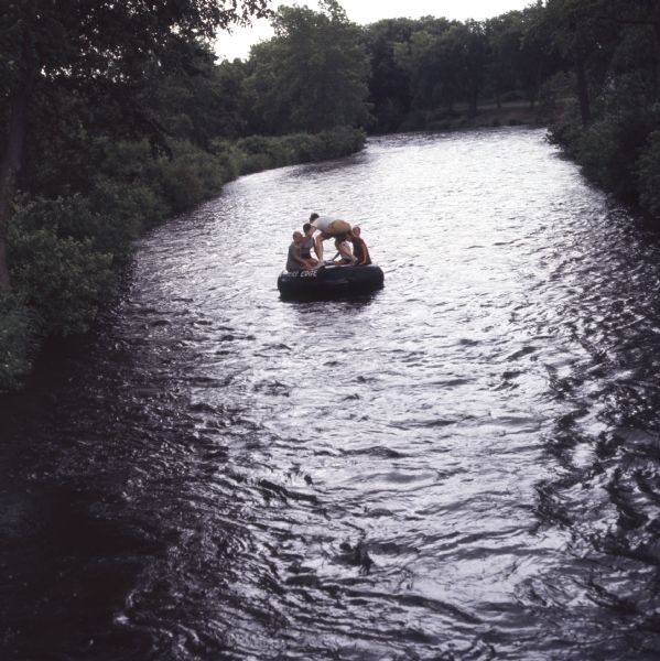 Elevated view down center of river towards a group of five people sitting and standing on a large rubber inner tube floating down Apple River.