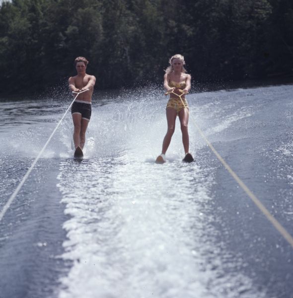 View from back of a boat of a young man and woman water skiing. The man is slalom skiing on the left, and the woman is using two skis on the right.
