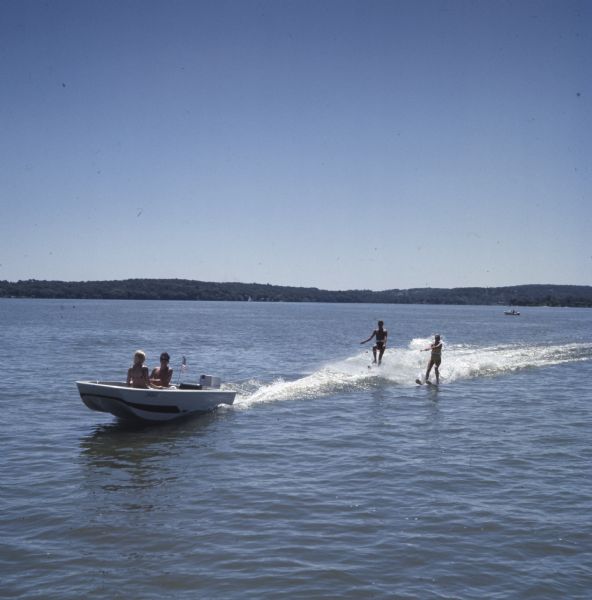 View across water towards a man and woman sitting in a motor boat, which is towing another man and woman water skiing behind it.