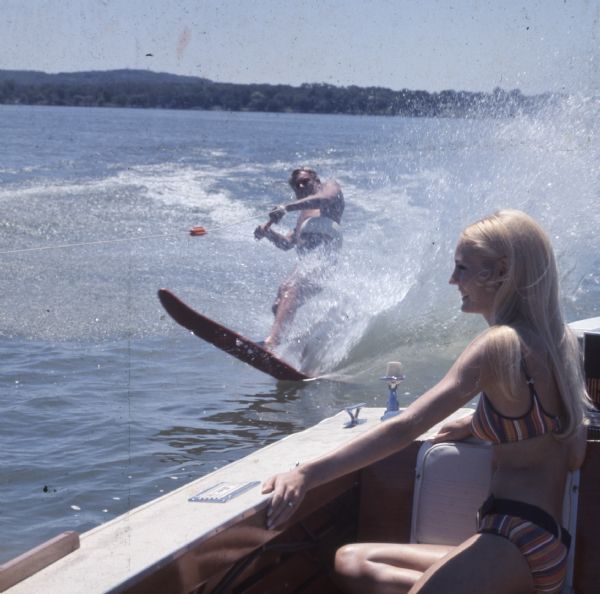 A young woman is sitting in the back of a motorboat watching a man slalom skiing just behind her.