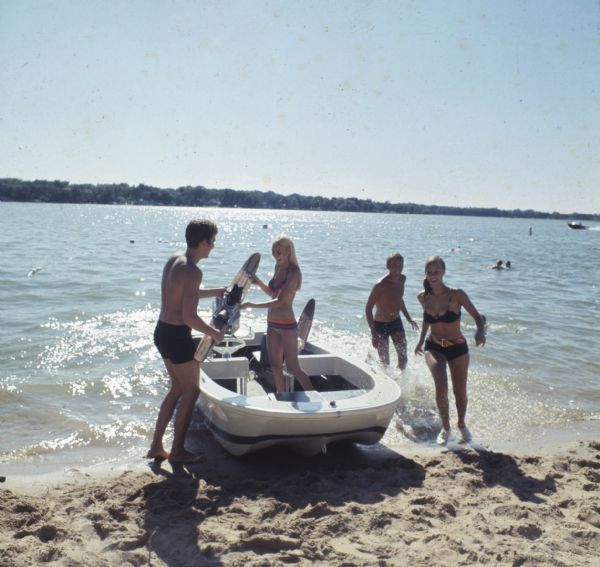 Four young men and women are standing in or around a motorboat beached on the sandy shoreline, with people swimming in the lake in the background. One woman is handing a man a slalom water ski.