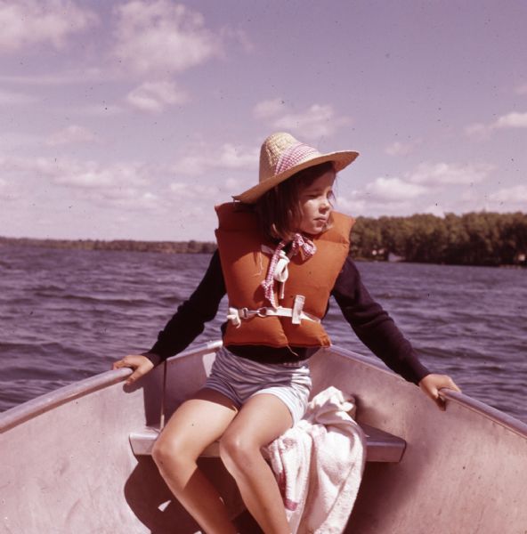 Close-up of a young girl sitting in the bow of a boat. She is wearing a life vest and a wide brimmed straw hat.