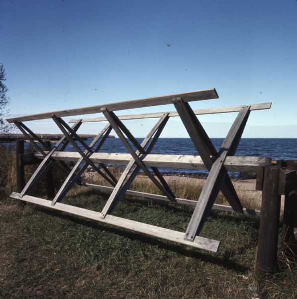 A wooden fishing net rack sitting on the grass near the shoreline of Lake Superior.