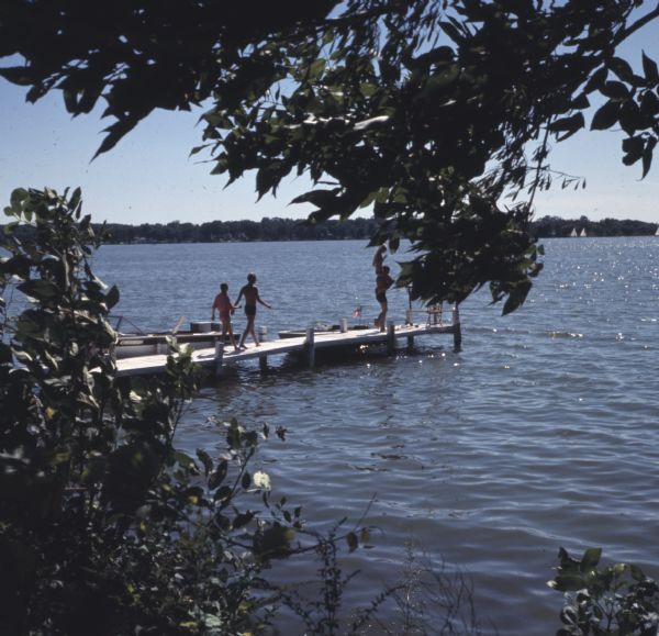 View from shoreline through the trees towards a man and a woman walking down a dock. At the end of the dock is a man is holding up a woman on his shoulders. Two motorboats are tied to the dock.