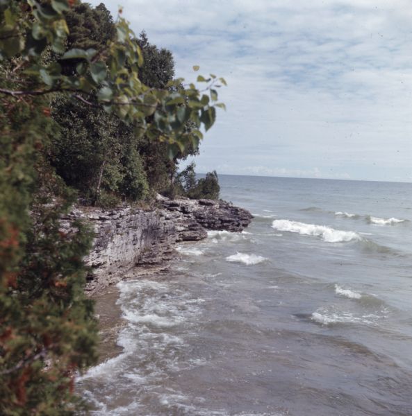 Elevated view of a rock outcropping along the tree-lined shoreline of Lake Michigan as waves are rolling in.