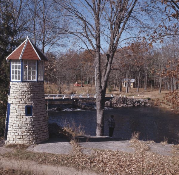 A little lighthouse on the shore of Little Wolf River. A person is standing on the shore near the lighthouse. In the background is a bridge spanning the river.