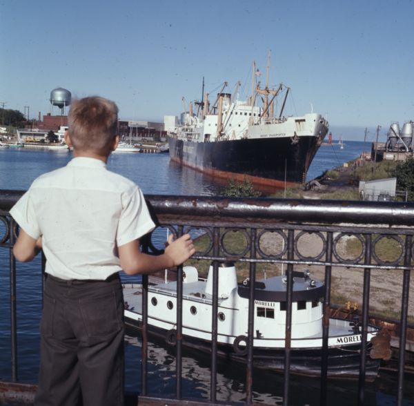 Rear view of a young boy leaning against a metal railing watching the ships in a harbor. A large cargo ship is in the harbor. A smaller boat, "Morelli," is below in the foreground. 
