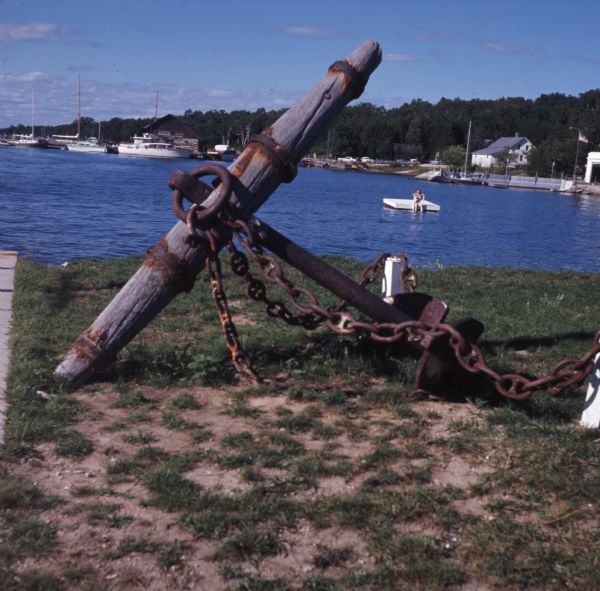 A wood and metal anchor is displayed on the shore of Gordon Lodge. A young girl is sitting on a floating raft in North Bay. Houses and boats are on the shoreline in the distance.