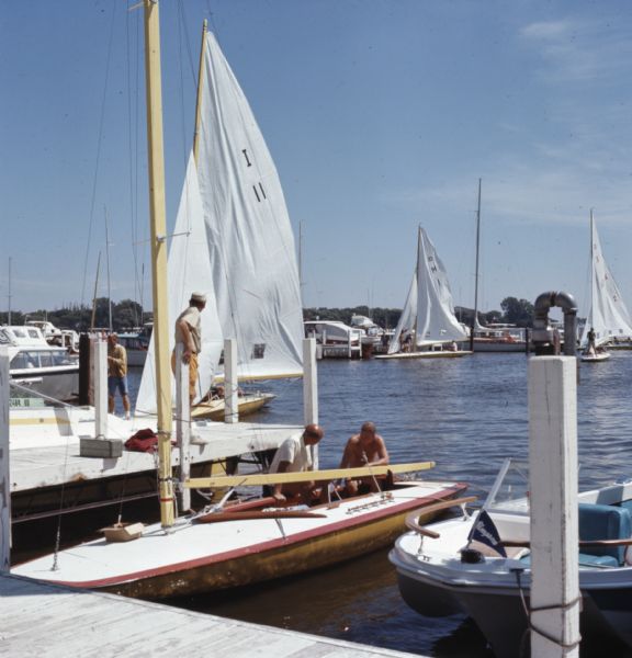 Sailboats are docked along a pier in a marina, along with some motorboats. Men are walking along the docks, and prepping their ships to sail.