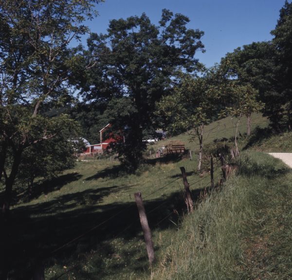 View along roadside toward a farm. A person is driving a tractor loaded with hay towards a red barn further down the road behind trees.