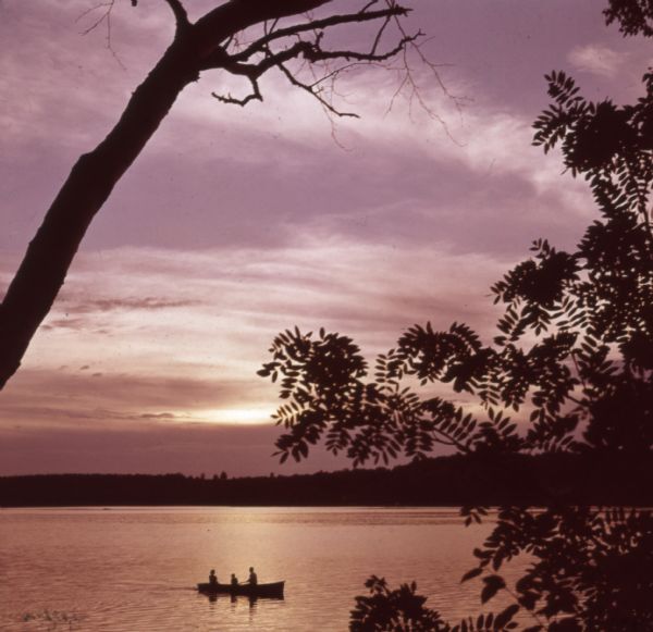 Three people are sitting in a canoe on a lake as the sun is setting.