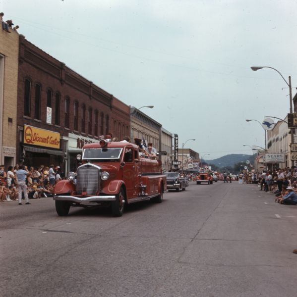 View down street towards a crowd of people, sitting or standing on the side of the street, or on top of the buildings, watching a parade. A firetruck from the Waterloo Fire Department is in the front of a line of cars and trucks.