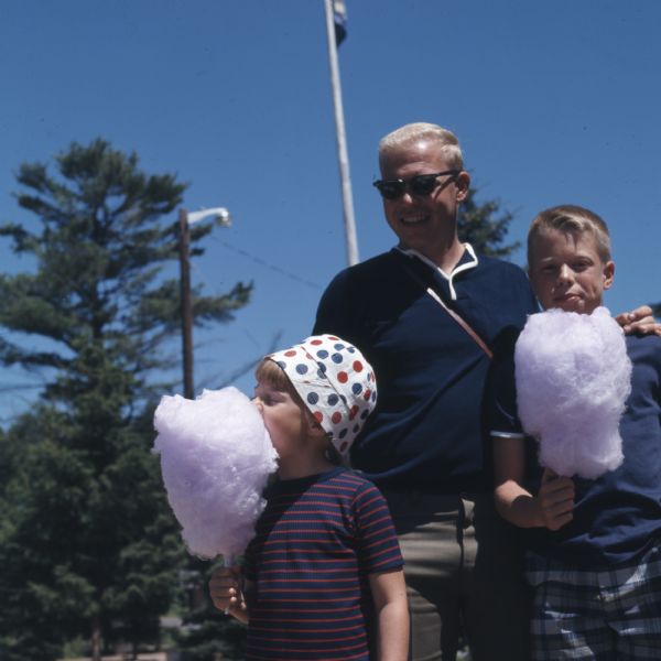 A father is standing outdoors next to his two children, who are eating pink cotton candy.