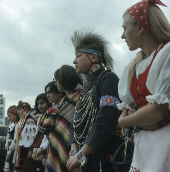 A group of young men and women are standing in a line outdoors wearing various types of ethnic clothing at the Holiday Folk Fair.