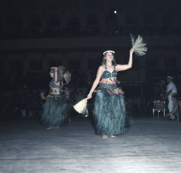 A group of women wearing green Polynesian dance costumes are dancing on a stage at the Holiday Folk Festival. A group of musicians are playing drums on the right, and a crowd of people are watching in the background.