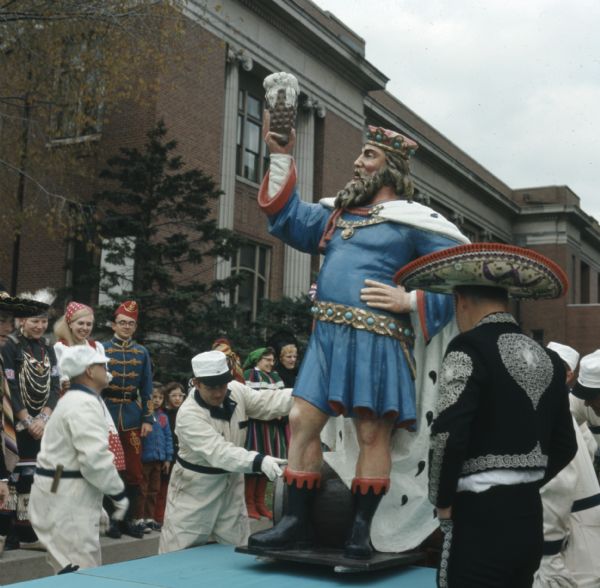 Men in white jumpsuits are steadying a statue of King Gambrinus on a parade float at the Holiday Folk Fair. A line of men and women in various ethnic clothing are watching from either side.