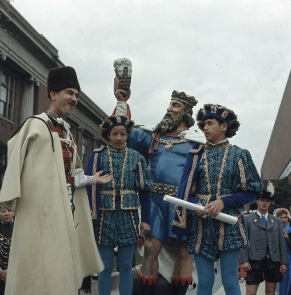 A man wearing Medieval European clothing is gesturing towards a statue of King Gambrinus on a parade float at the Holiday Folk Fair. Two boys, in matching Medieval European outfits, are standing on either side of the statue. One boy is holding a scroll. Other people in various ethnic dress are standing around the float.