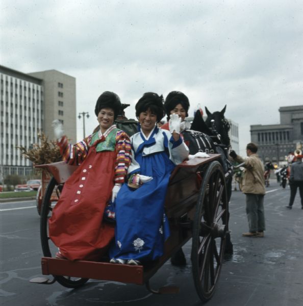 Three women in traditional Korean hanbok are riding in the back of a horse-drawn wagon and waving to the photographer.