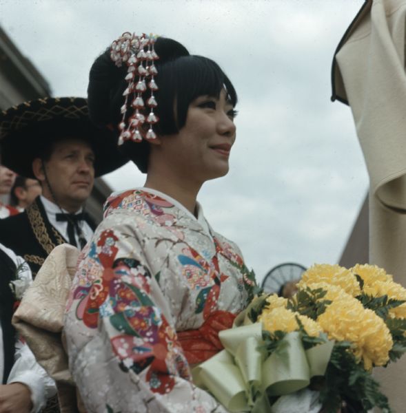 Irene Janokuchi, the official Holiday Folk Fair hostess, is wearing a white and red Japanese kimono and holding a bouquet of yellow chrysanthemums.