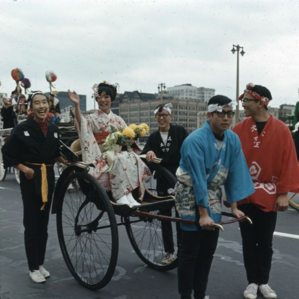 Irene Janokuchi, the official Holiday Folk Fair hostess, is riding in a rickshaw in the parade. She is wearing a red and white kimono and holding a bouquet of yellow chrysanthemums. Four young men, wearing a mixture of modern and traditional Japanese clothing, are standing nearby or pulling the rickshaw.