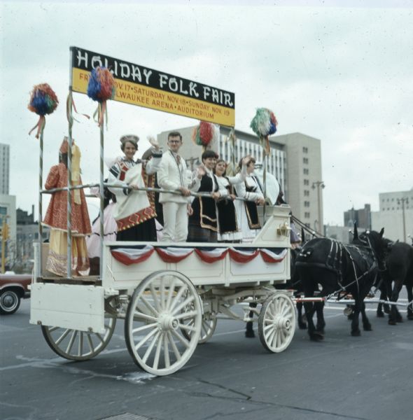 Women and men in traditional European and Middle Eastern clothing are waving from a white carriage pulled by four horses. A sign on the carriage reads: "Holiday Folk Fair: Firday, Nov. 17, Saturday Nov. 18, Sunday Nov. 19, Milwaukee Arena, Auditorium."