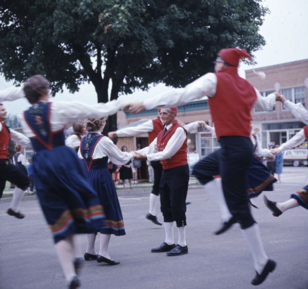 A group of people wearing costumes are performing a Norwegian dance at the Song of Norway Festival. A group of people are dancing in a round, while a couple is dancing in the middle of the circle.