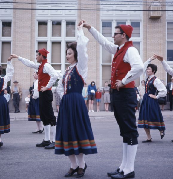 A troupe of dancers are performing a Norwegian dance in pairs at the Song of Norway Festival. The men are wearing red hats, red vests over a white shirt, and black breeches. The women are wearing blue dresses with floral patterns on the bodice over a white shirt.