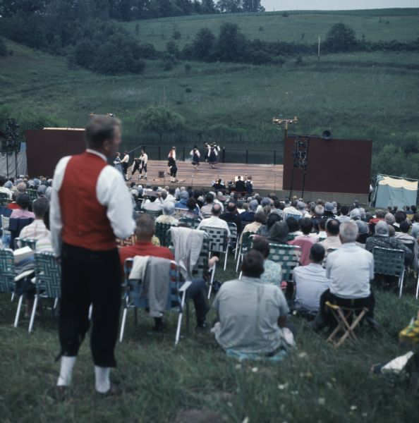 View down hill towards actors performing the musical "Song of Norway" on an outdoor stage at the Song of Norway Festival. People are sitting on blankets or in lawn chairs on the grassy hill to watch the production.