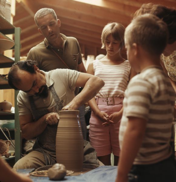A small audience of children and adults are watching a ceramicist throwing a vase on a pottery wheel. Pottery is sitting on shelving on the left.
