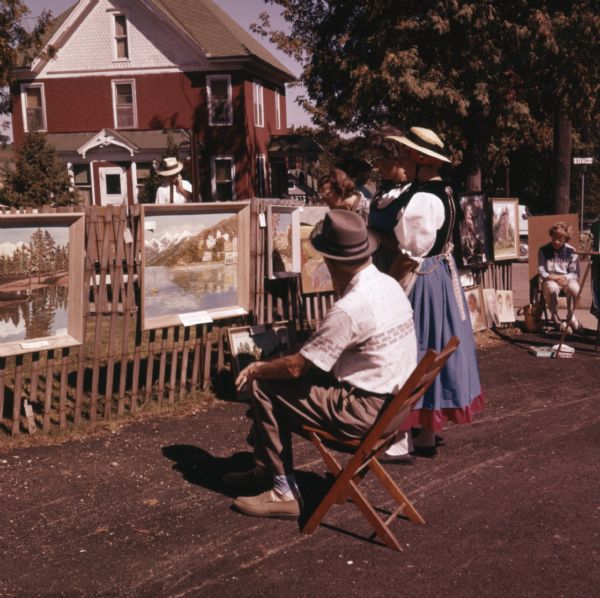 Artists and vendors are sitting in wooden chairs next to displays of paintings for sale. Two women, dressed in traditional Swiss dresses, are looking at the paintings.