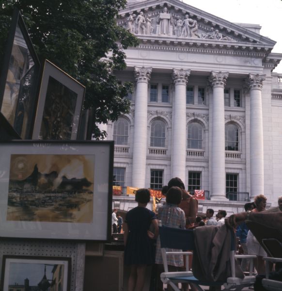 Crowds of people around the Wisconsin State Capitol at an art fair. On the left, paintings are displayed for sale.