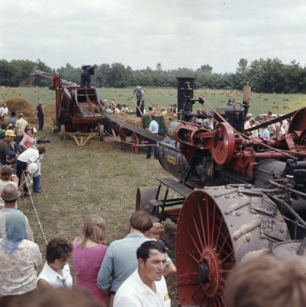 Elevated view of a group of men demonstrating the use of a tractor belt powering a threshing machine. A crowd of people are watching from the sidelines.