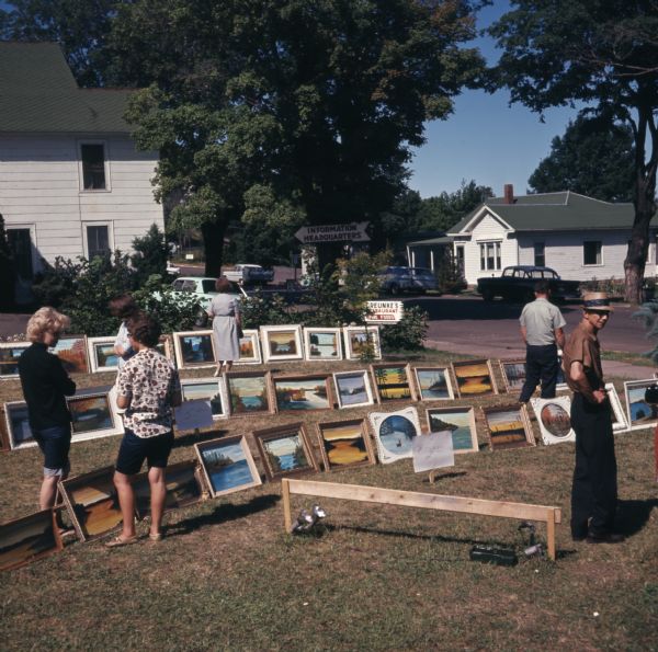 Three rows of paintings are displayed outdoors on a lawn. Men and women are walking among the rows looking at the pieces for sale.