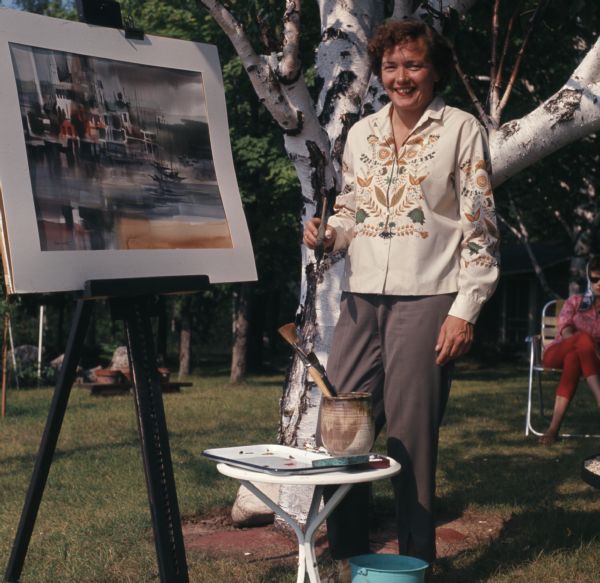 Full-length portrait of Doris White standing outdoors. She is holding a paintbrush in her right hand. A small table is in front of her, holding brushes and a paint palette. She is standing next to one of her paintings, displayed on an easel. 