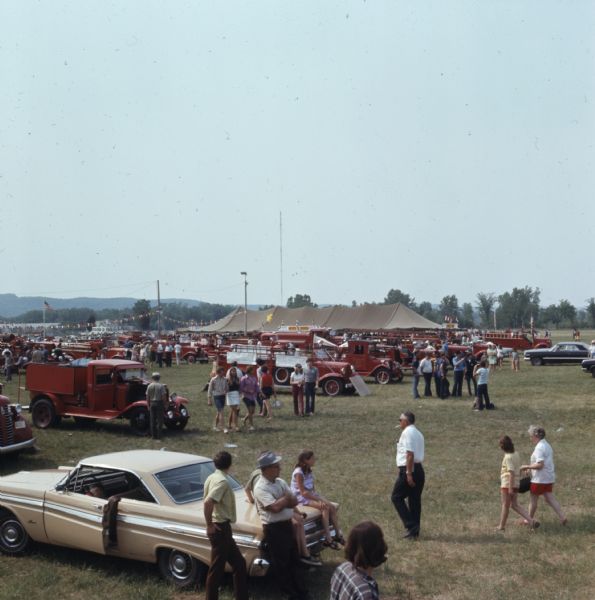 Elevated view of fire trucks parked in rows at a fairgrounds, with a crowd of people walking between them. In the background is a large tent advertising cold beer.