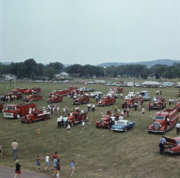 Elevated view of a crowd walking among fire trucks parked in rows at an outdoor fairgrounds.