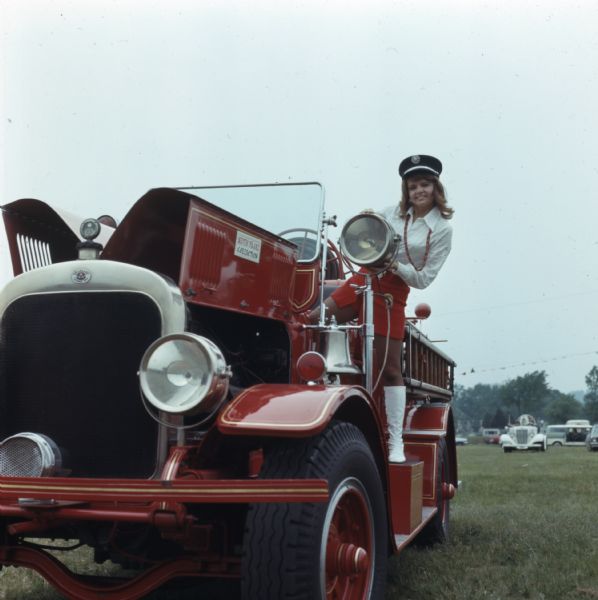 A woman wearing a fire department officers cap, with a white shirt, red skirt, and tall white boots is posing standing on a fire truck, holding onto one of the headlights. A metal sign on the truck reads: "Keith Franz Collection." 