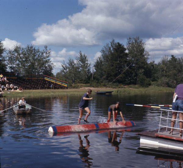 Two young women are competing in logrolling. People watch from bleachers on a lawn in the background. One person is sitting and watching from a rowboat, and another person is standing on a floating raft on the right.