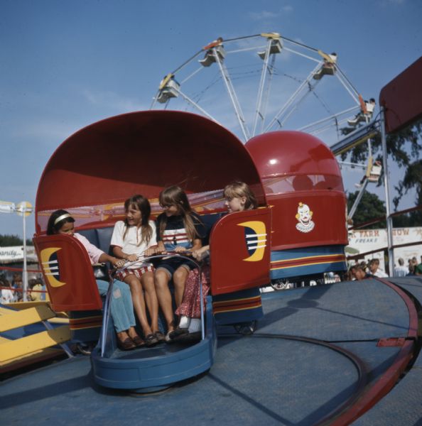 Four girls are sitting in a tilt-a-whirl amusement ride. In the background people are riding on a Ferris wheel.