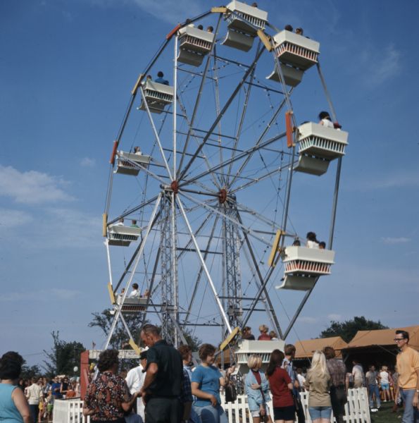 People are riding on a Ferris wheel above a crowd of people walking around a fairgrounds.