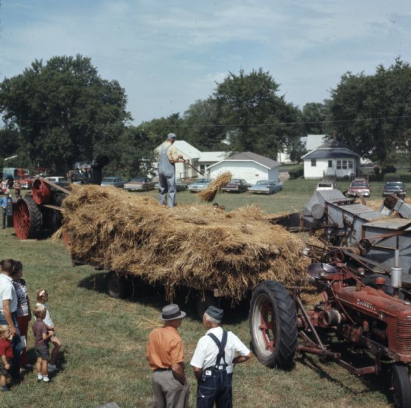 Elevated view of a man standing on a hay wagon, lifting hay with a pitchfork to load it onto a steam engine powered thresher. A group of people are watching the demonstration.