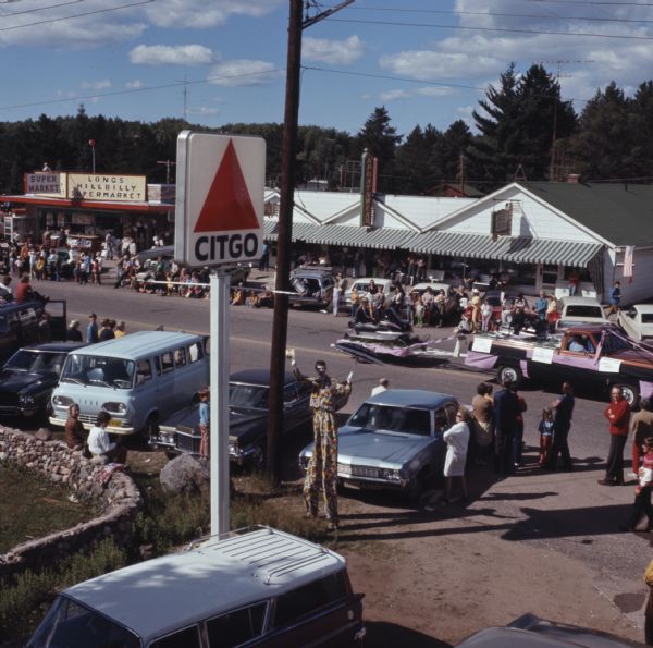 Elevated view of a crowd of people standing or sitting on the side of a road. Many of them are sitting on the back of their cars. A truck, decorated in pink and black cloth and streamers, is pulling a trailer carrying a woman sitting on a snowmobile. Near the Citgo sign in the foreground is a clown on stilts holding up an umbrella and tipping his hat.