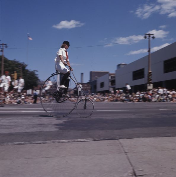 Side view of a woman riding a penny-farthing down the street during the Old Milwaukee Days annual circus parade. A crowd of people are sitting or standing along the sidewalk in the background.
