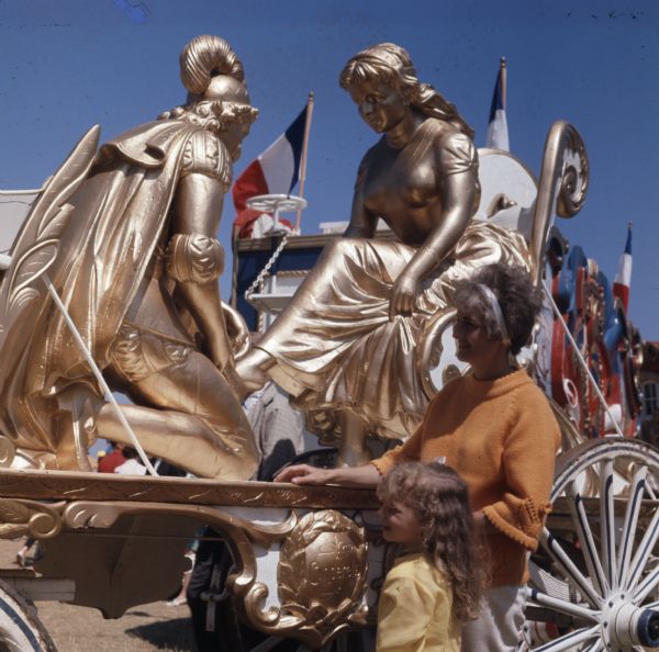 A mother and a daughter are looking at a gold colored statue on a circus wagon during Old Milwaukee Days. The statue depicts Cinderella sitting in a chair while the prince is fitting the glass slipper on her foot. The wagon in the background is decorated in four French flags and is painted in blue, white, and red.