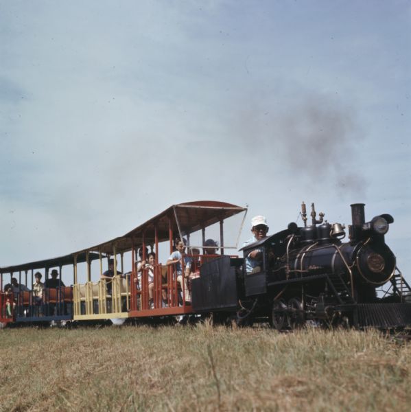A man, wearing a white cap reading "Strum Steam Engine Days," is driving a small, vintage narrow gauge train. People are riding in the train cars behind him.