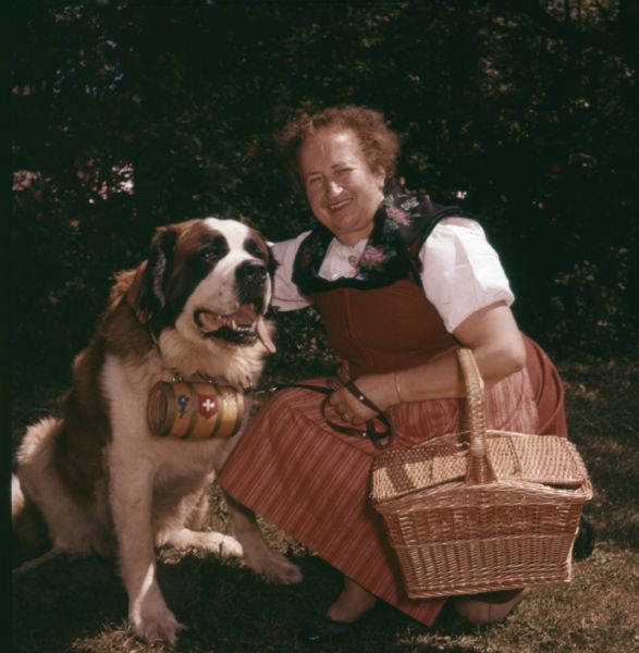 A woman carrying a basket over her arm and dressed in a Swiss dress is kneeling and posing with her Saint Bernard. A small wooden keg decorated with the Swiss flag is hanging from the dog's collar. 
