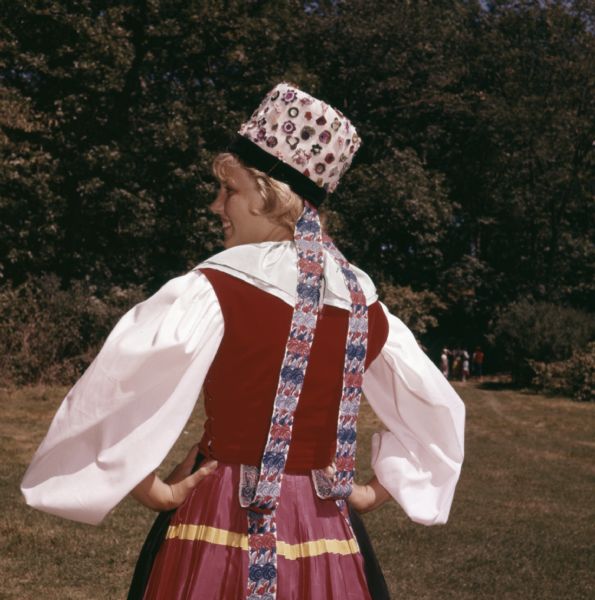 A woman with her back turned is posing outdoors and showing the back of her Swiss costume. Her hat is decorated with pins.